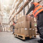 Two men moving boxes on a pallet inside a warehouse setting.