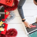 A woman using a laptop to shop online sitting next to wrapped presents.