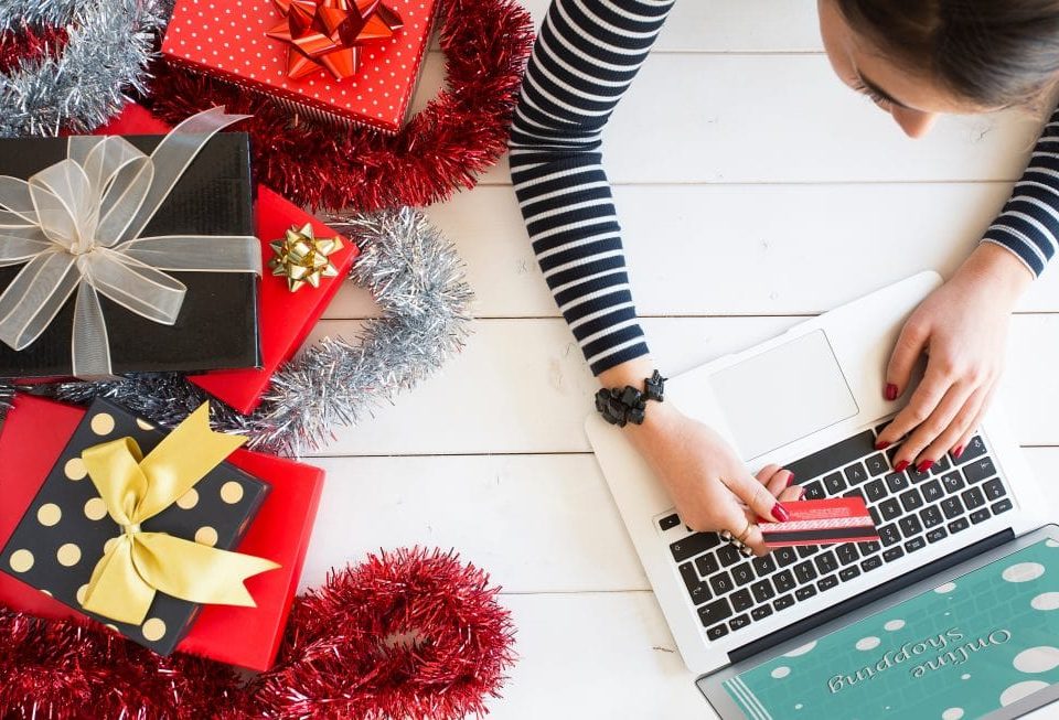 A woman using a laptop to shop online sitting next to wrapped presents.