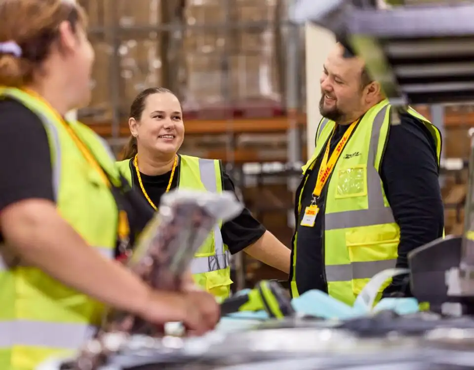 Three DHL employees are having a conversation in a warehouse.