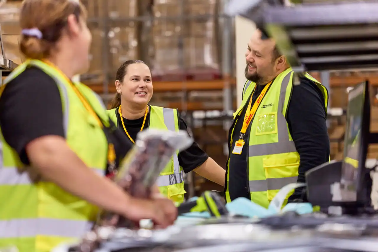 Three DHL employees are having a conversation in a warehouse.